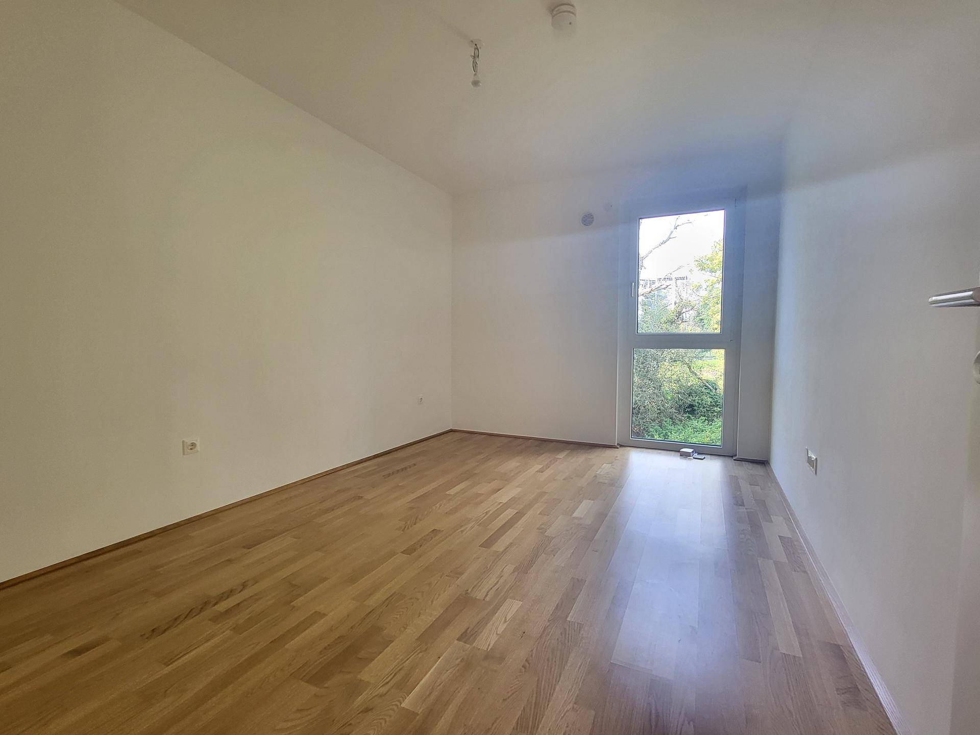 Helles Zimmer mit Holzparkett und großem Fenster mit Blick ins Grüne.