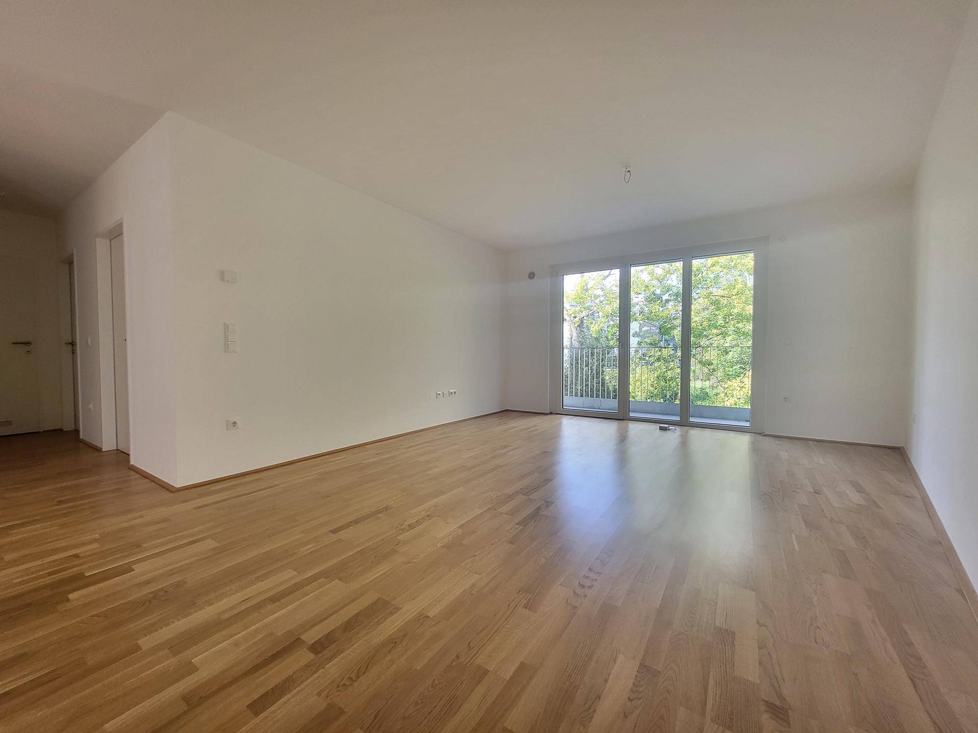 Weitläufiger Wohn-Essbereich mit hellem Holzfußboden und großen Fenstern zum Balkon.