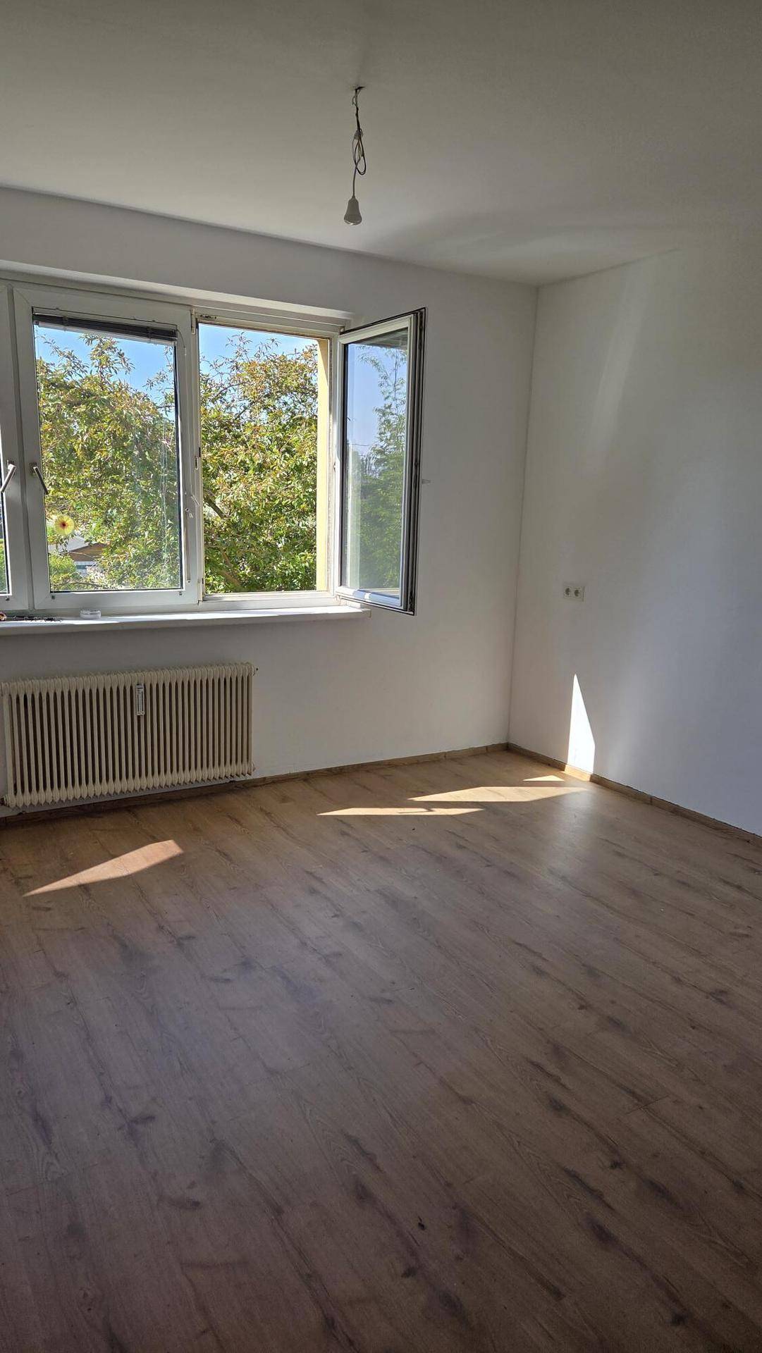 Lichtdurchflutetes Zimmer mit Holzboden und großem Fenster, das einen Blick ins Grüne bietet.