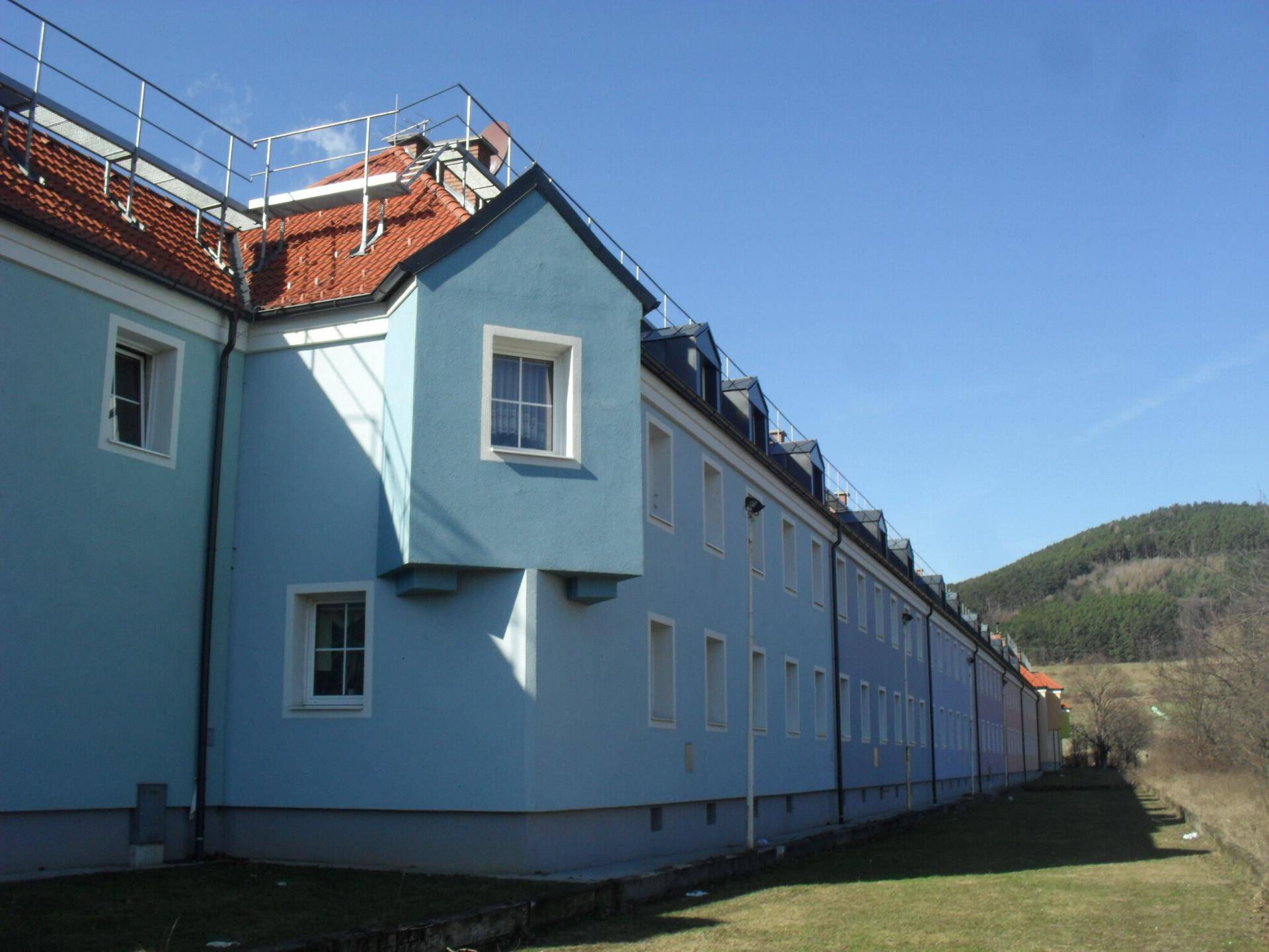 Lange Gebäudefassade in Hellblau mit Dachgauben und angrenzender Grünfläche unter blauem Himmel.