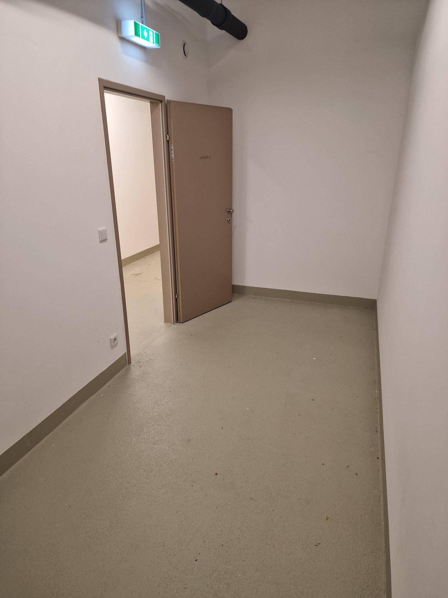 Leerer Lagerraum mit weißen Wänden und hellem Boden, eine offene Tür führt zu einem weiteren Raum.