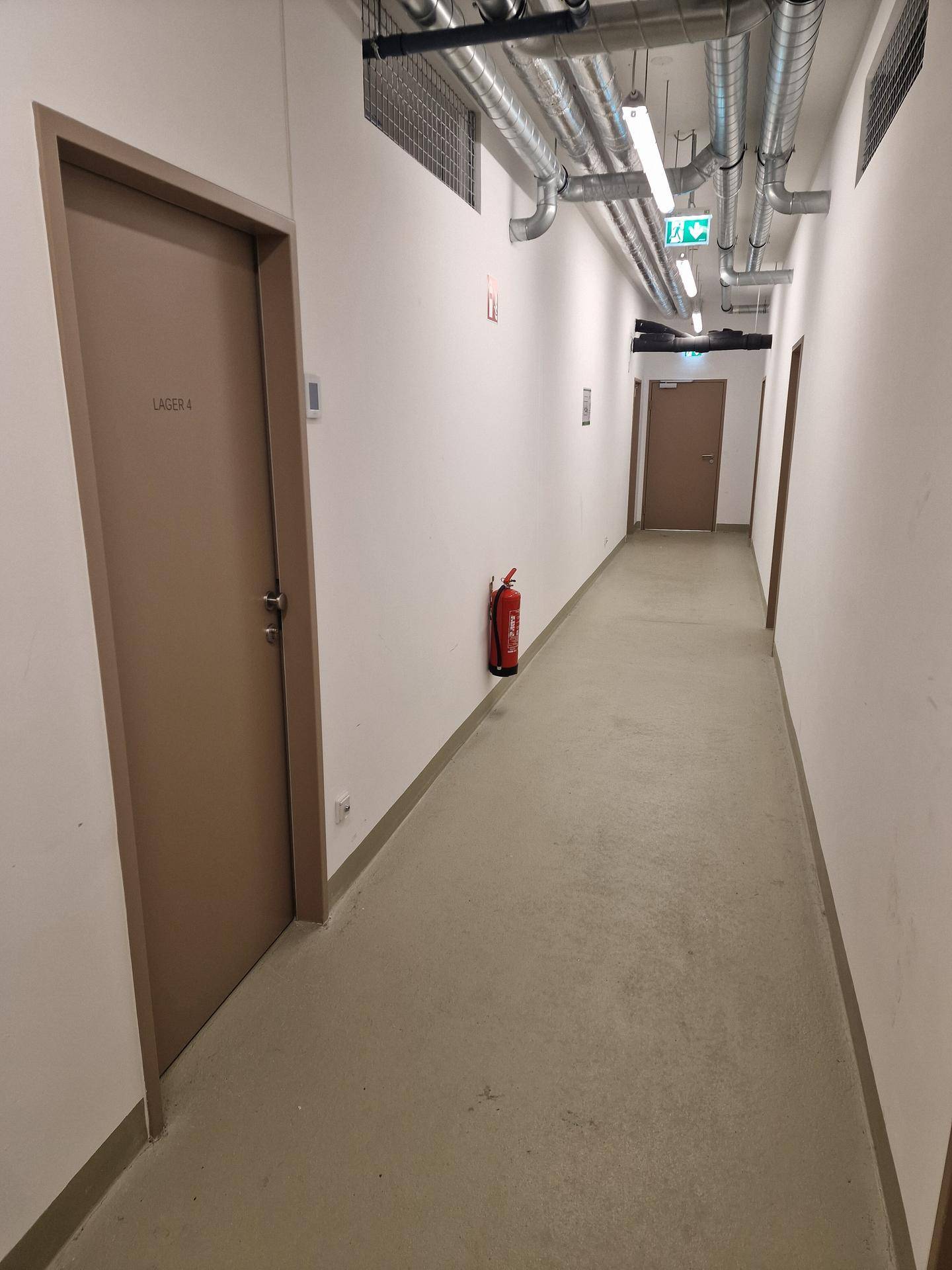Langer Flur mit mehreren Türen zu Lagerräumen, sichtbaren Leitungen an der Decke und einem Feuerlöscher.