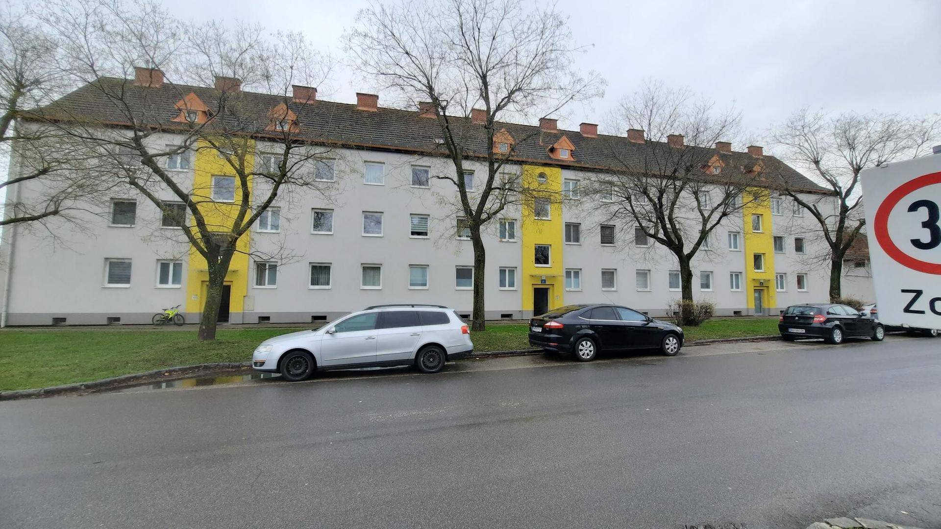 Langes Wohngebäude mit gelben und grauen Fassadenabschnitten entlang einer Straße mit parkenden Autos.