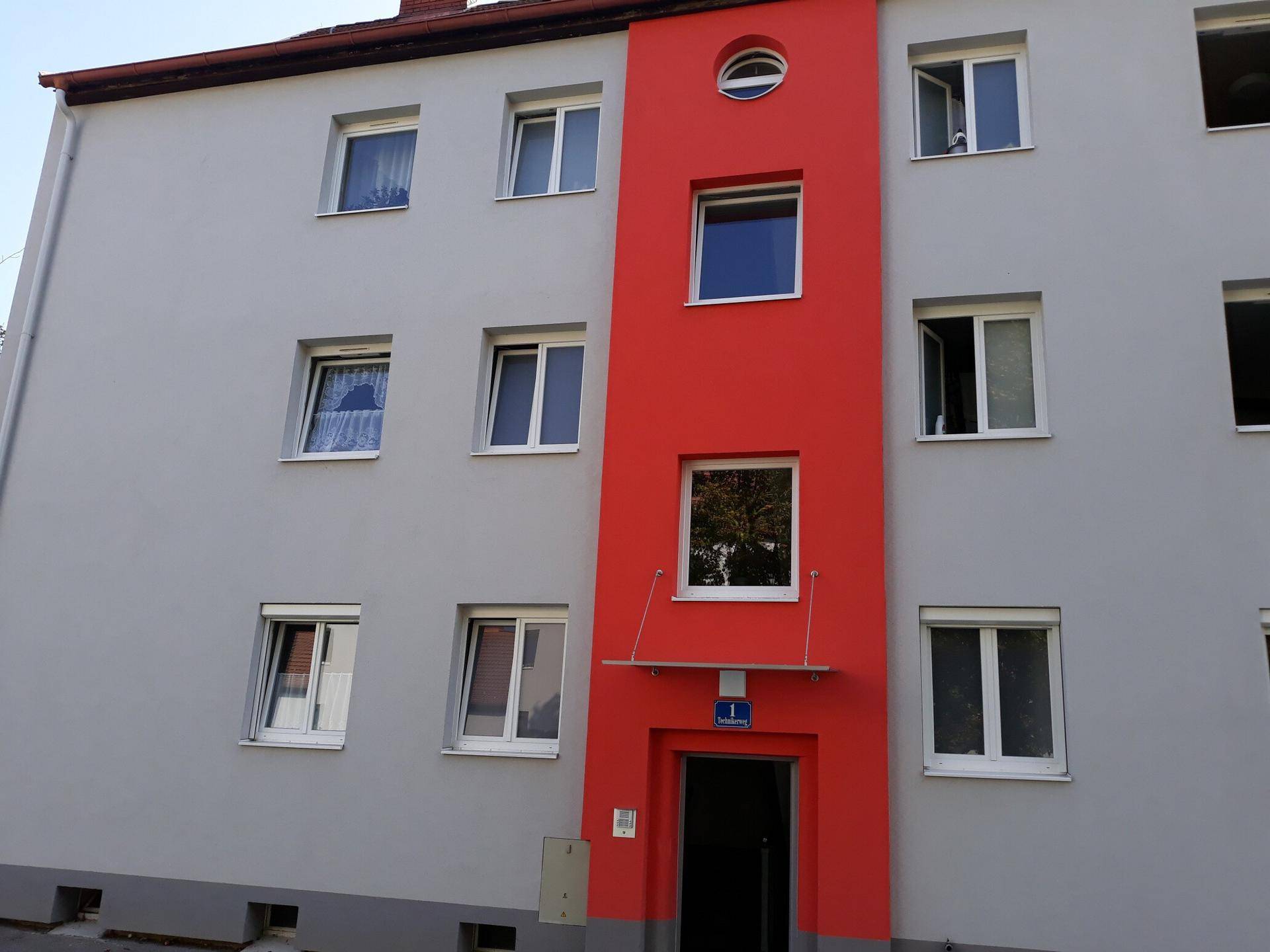 Fassade eines Wohngebäudes mit markantem roten Akzent und grauen Wänden am Eingangsbereich.