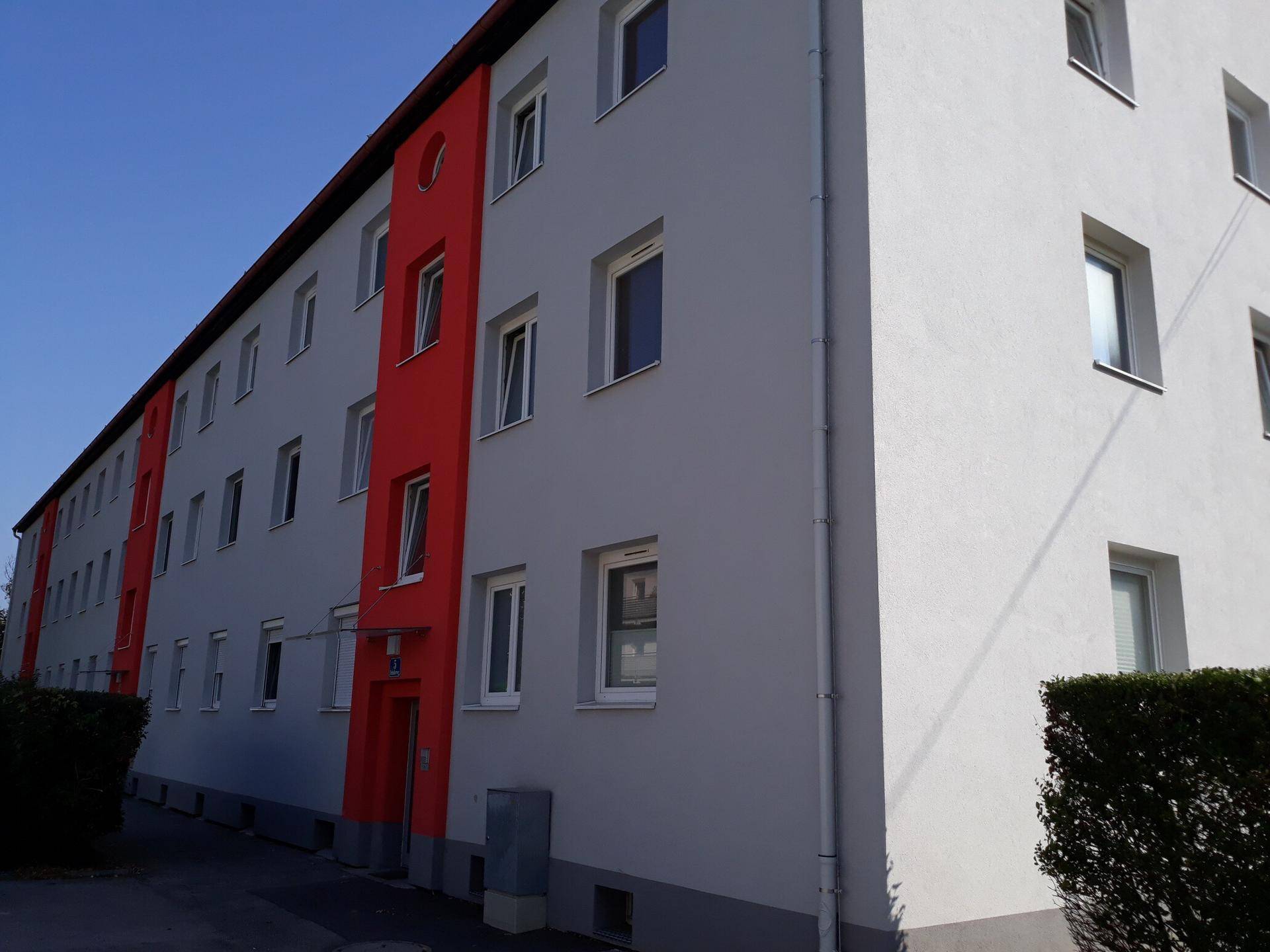 Seitenansicht eines Mehrfamilienhauses mit roter und grauer Fassade und gepflegten Hecken.