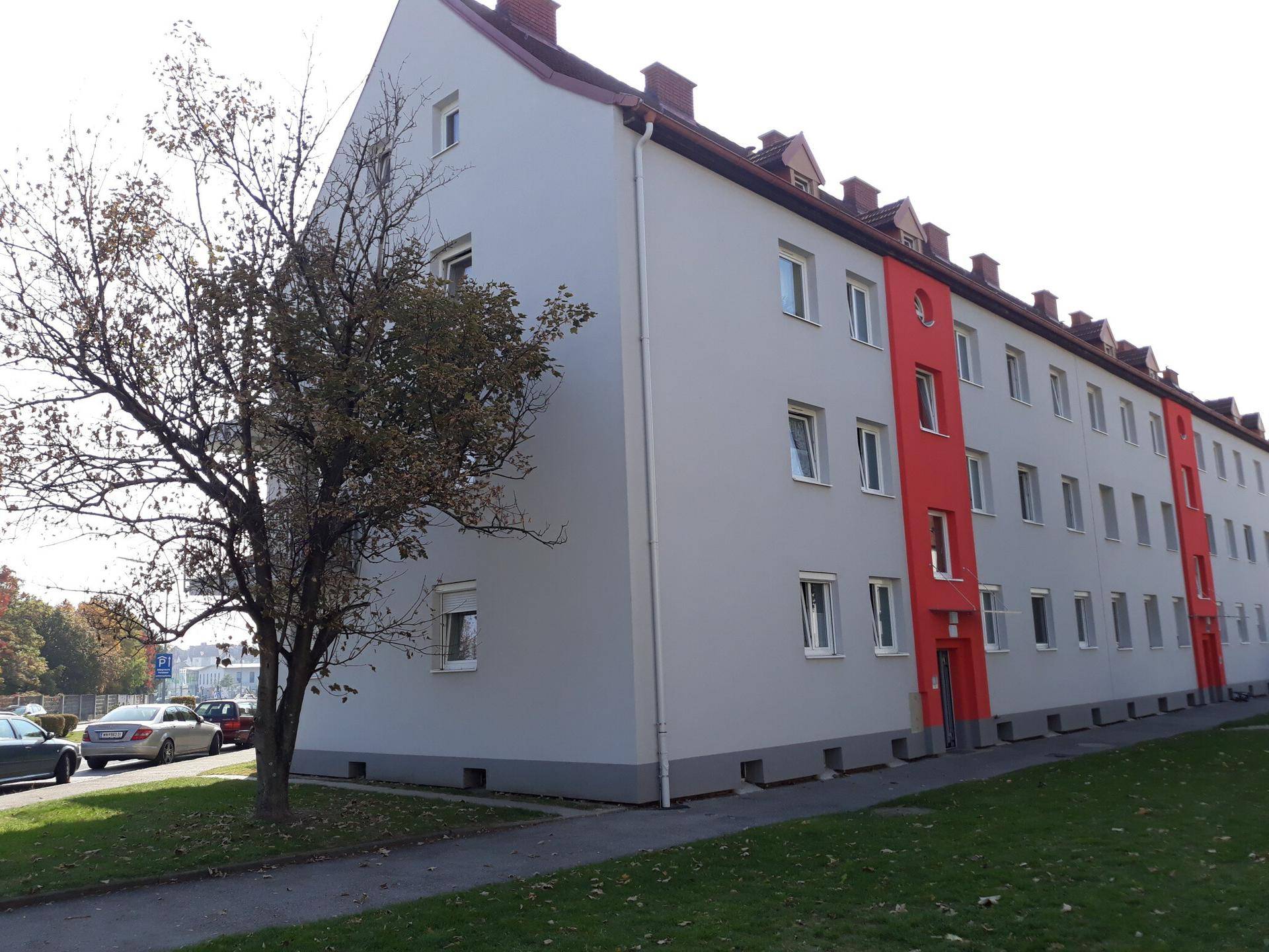 Wohngebäude mit roter und grauer Fassade, umgeben von Rasenflächen und einem Baum unter blauem Himmel.