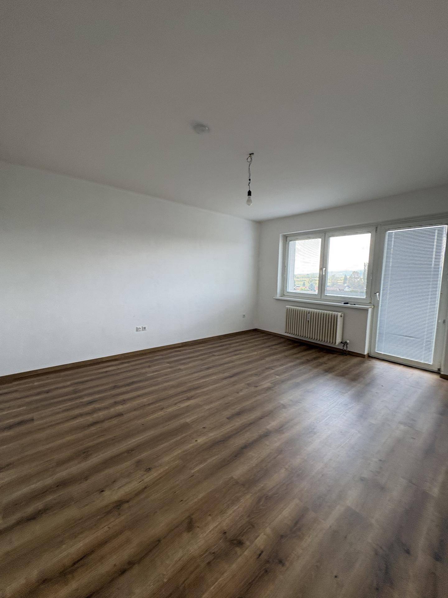 Helles, leeres Zimmer mit Laminatboden, weißen Wänden und Zugang zu einem Balkon oder einer Terrasse.