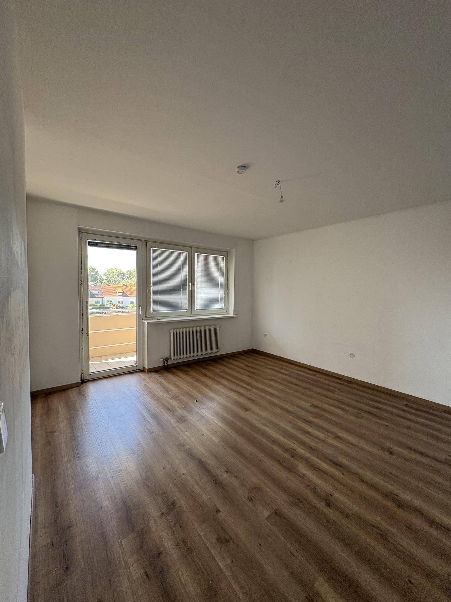 Leeres Zimmer mit Laminatboden, großen Fenstern und Zugang zu einem Balkon.