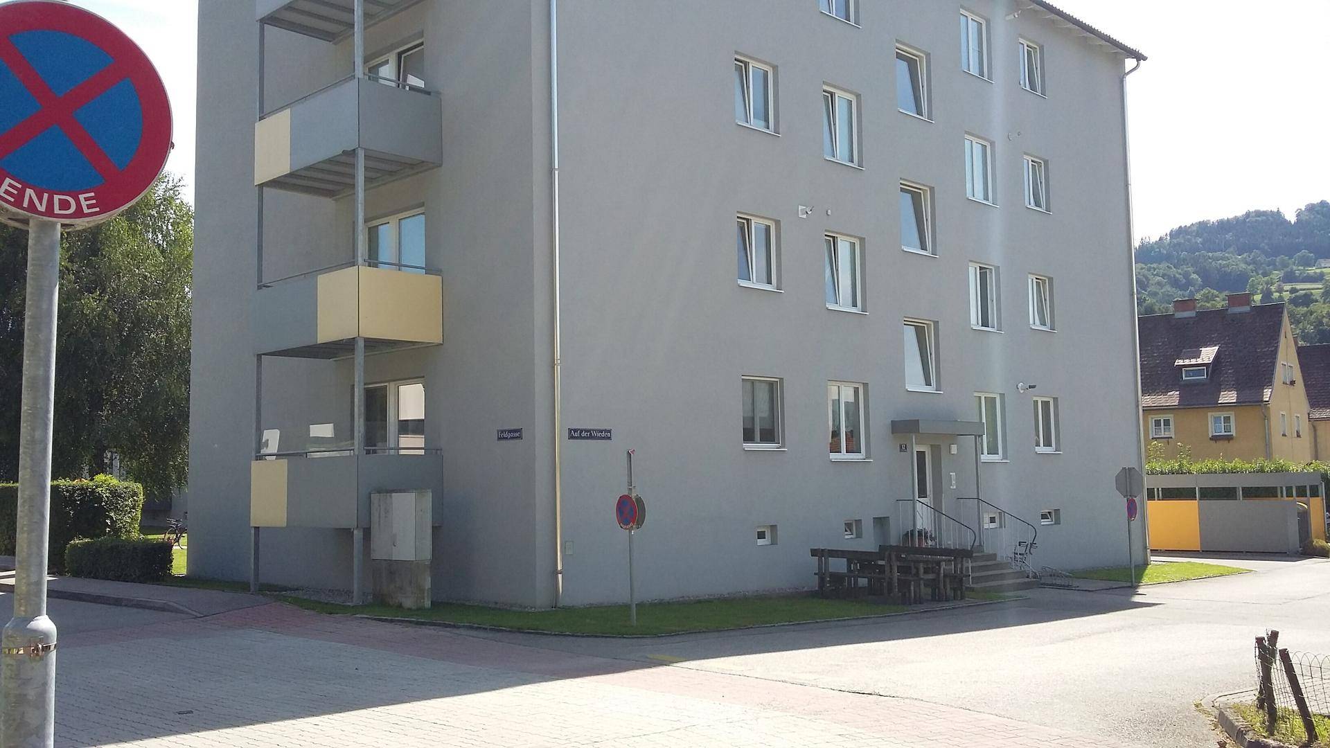 Eckansicht eines grauen Wohngebäudes an einer Straße mit einem Verkehrsschild und Hügeln im Hintergrund.