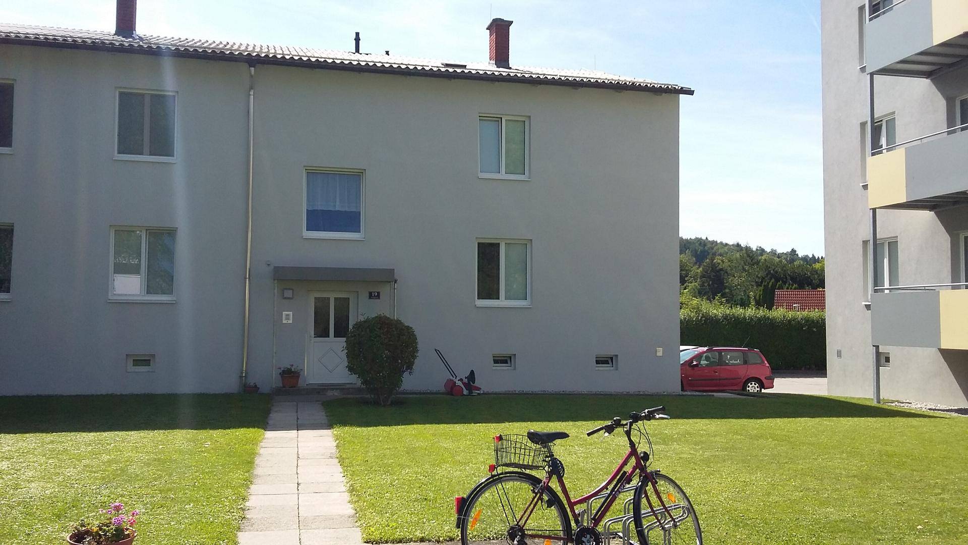 Rückseite eines grauen Wohnhauses mit Eingangsbereich und einer grünen Rasenfläche an einem sonnigen Tag.