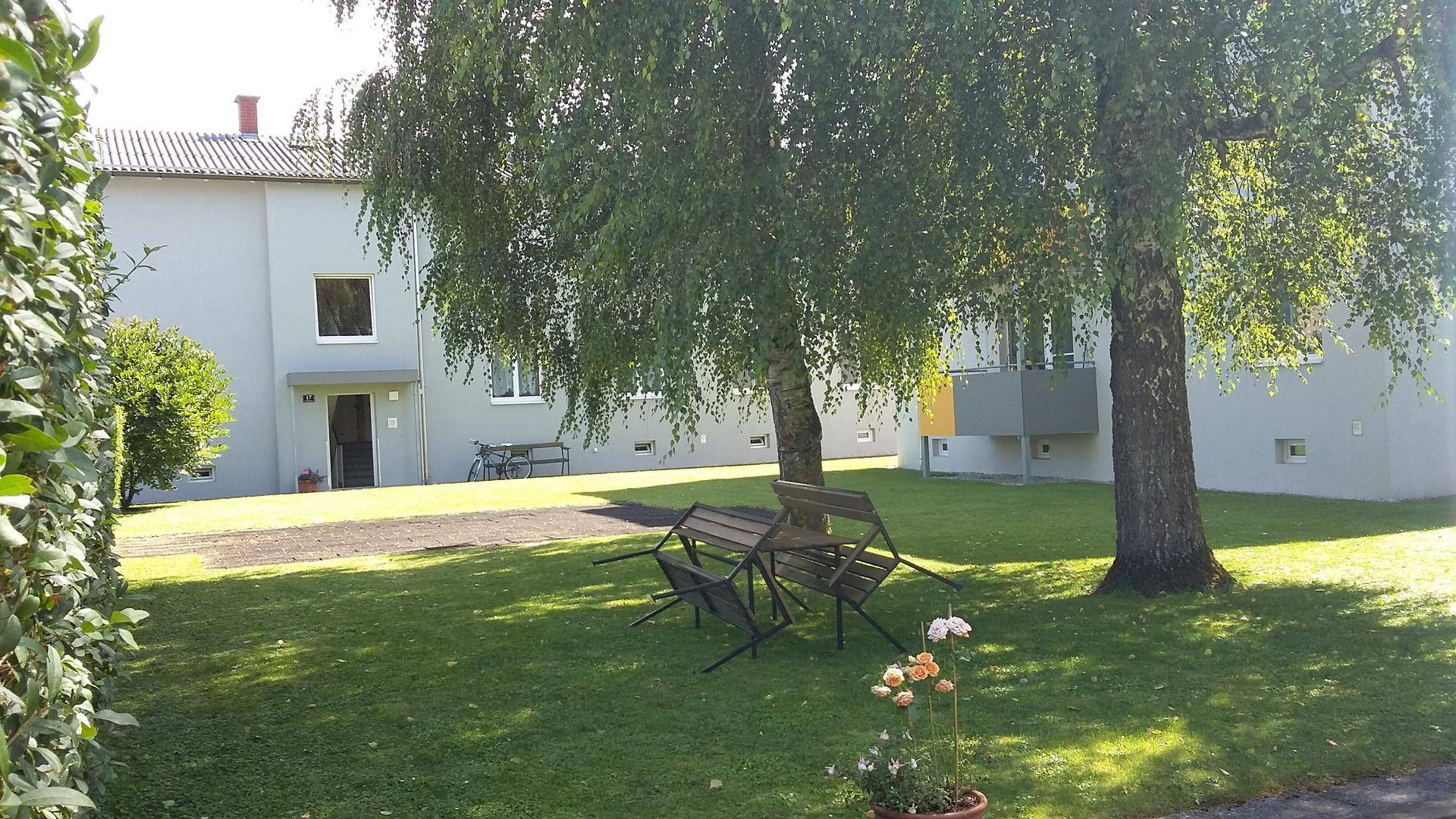 Gepflegte Grünfläche mit Bäumen und einfachen Sitzgelegenheiten vor einem modernen Wohngebäude.