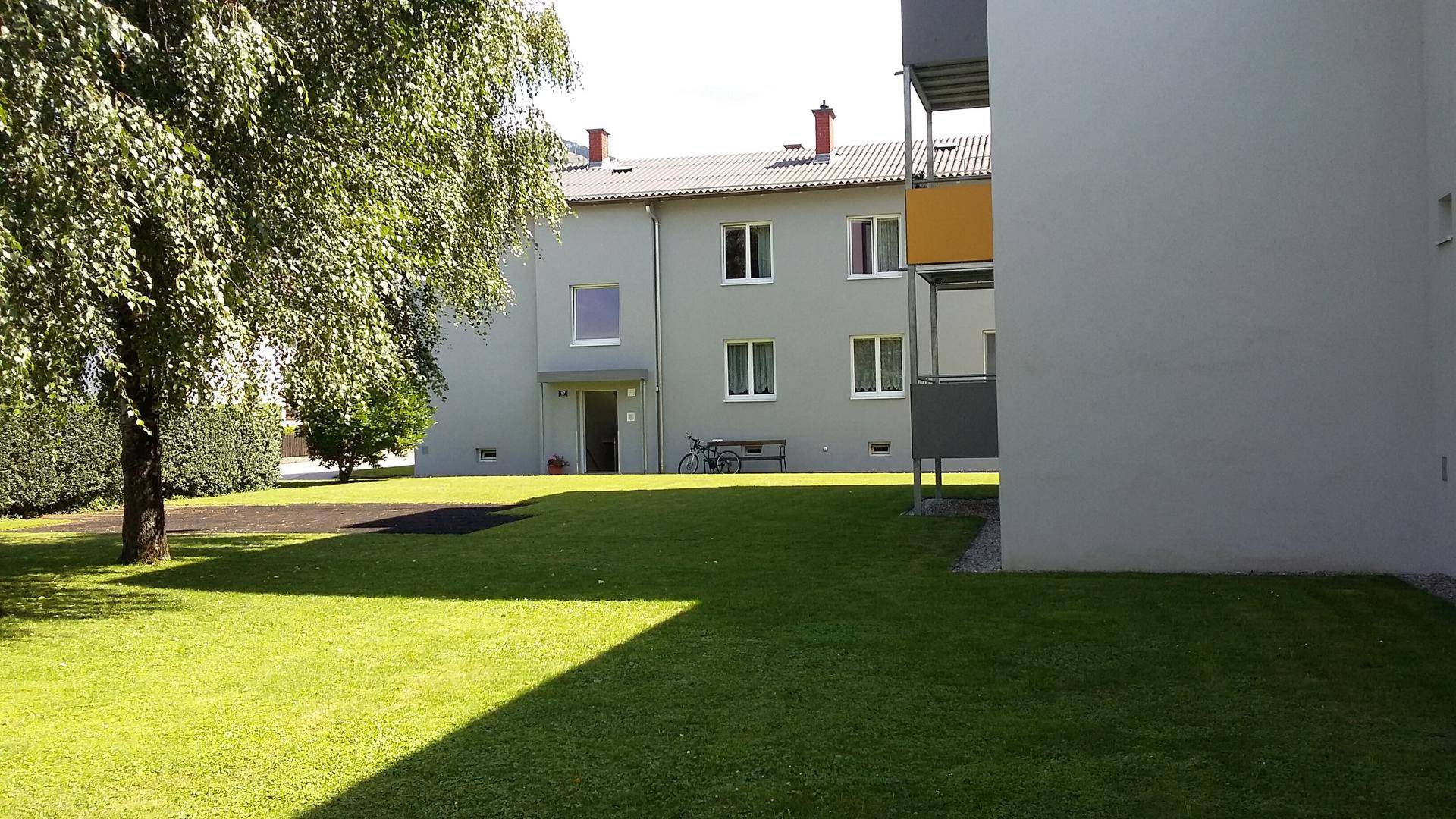 Wohngebäude mit einem großen Baum und einer weitläufigen Rasenfläche im Vordergrund bei Sonnenschein.