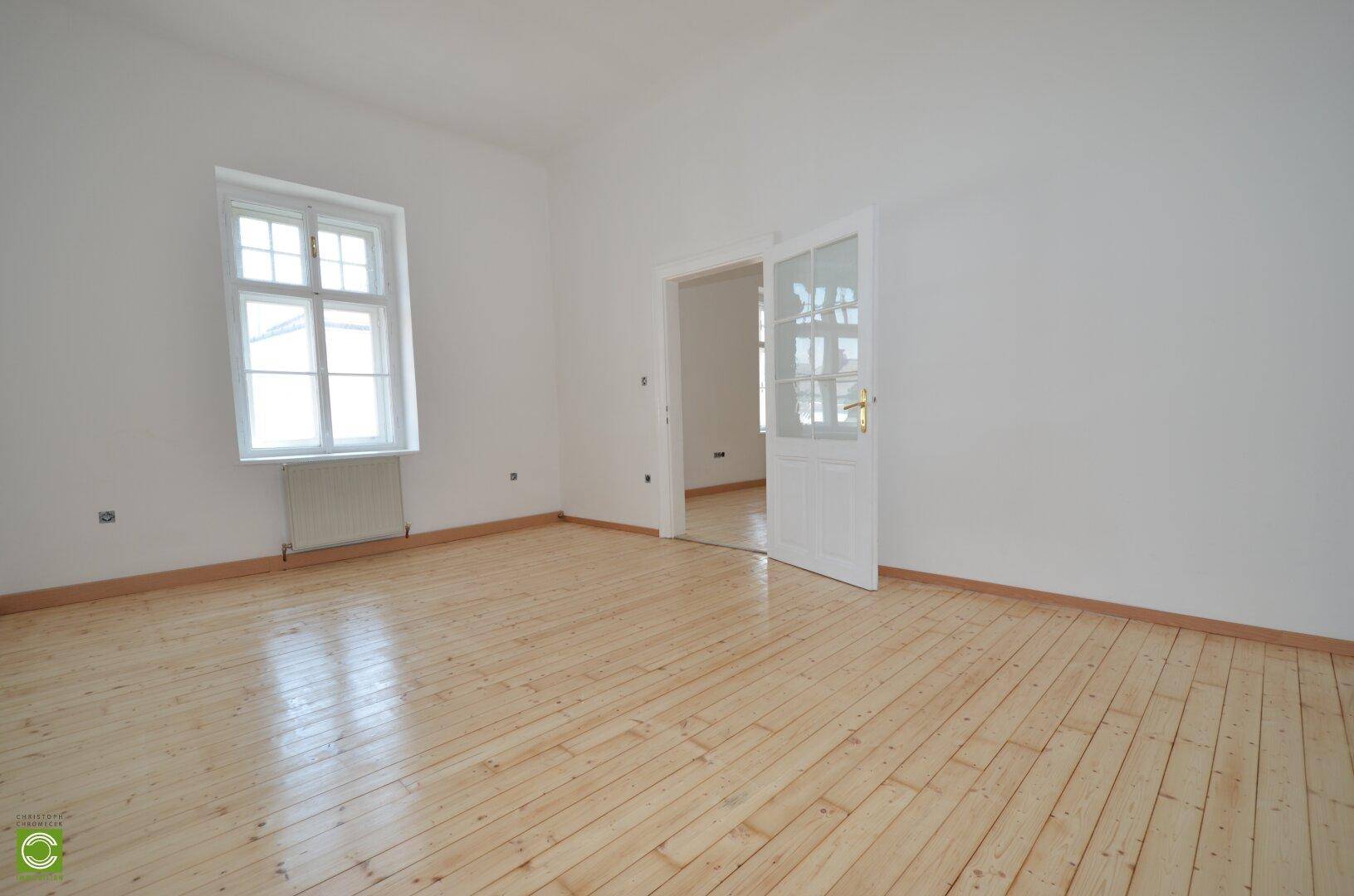 Heller Raum mit Holzdielenboden und einer Glastür, die zu einem weiteren Zimmer führt.