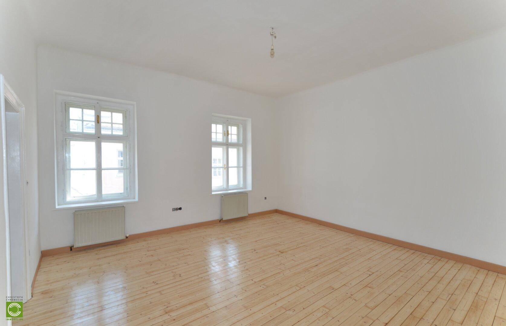 Geräumiger, heller Raum mit Holzdielenboden, weißen Wänden und zwei Fenstern.
