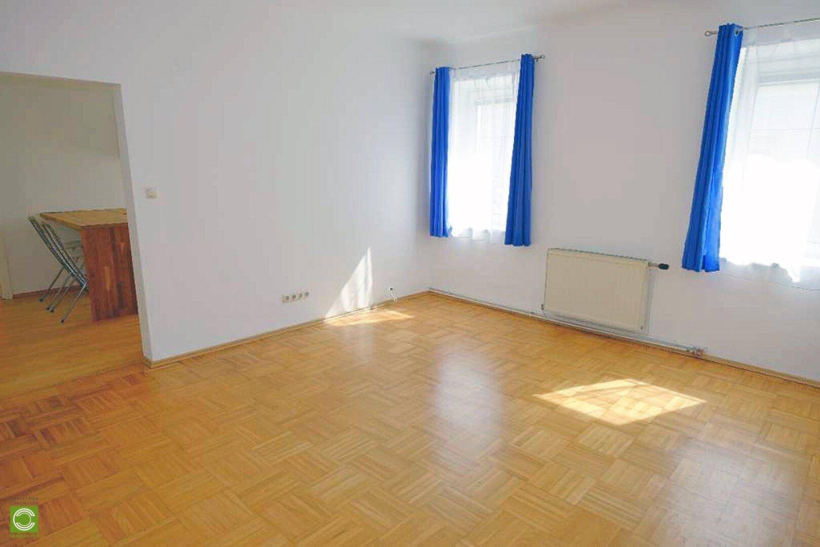 Helles, leeres Wohnzimmer mit Parkettboden und blauen Vorhängen, bereit zur Einrichtung.