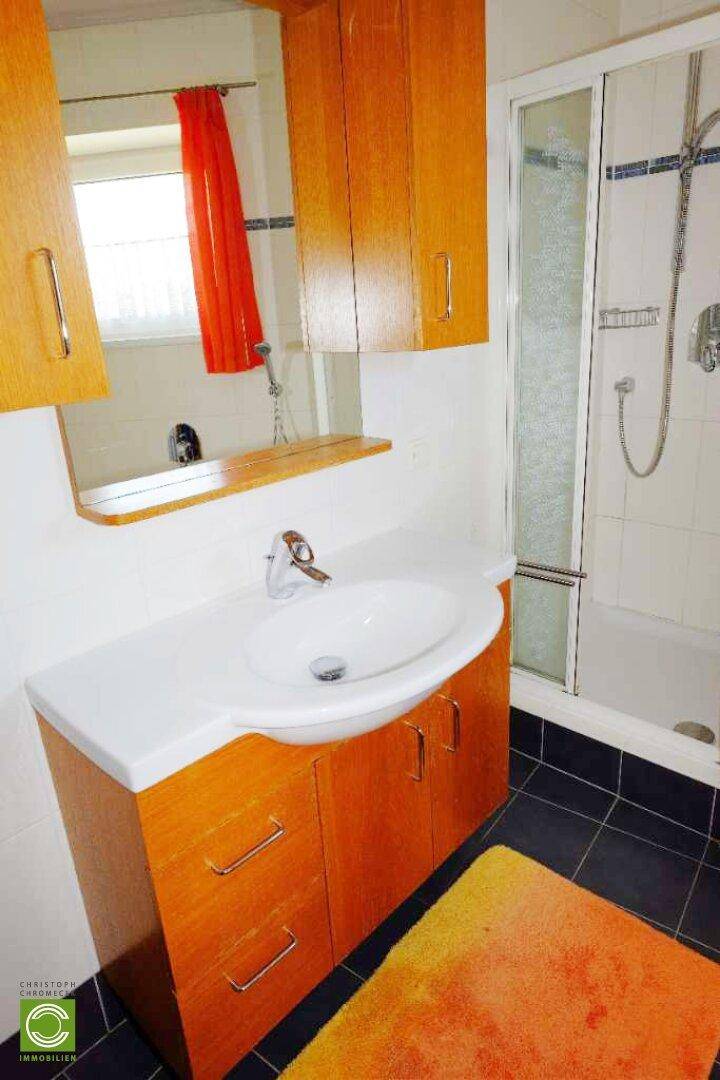 Badezimmer mit Waschbecken, Dusche und Holzschränken, ausgestattet mit Spiegel und Fenster.