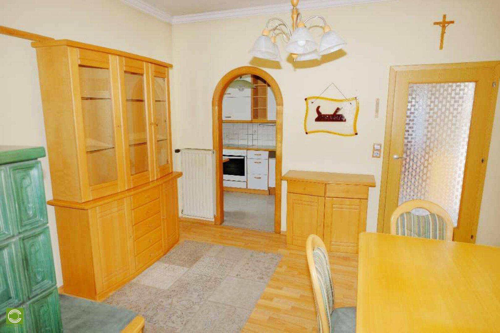 Essbereich mit traditionellem Kachelofen und Holzmöbeln, mit Blick in die angrenzende Küche.
