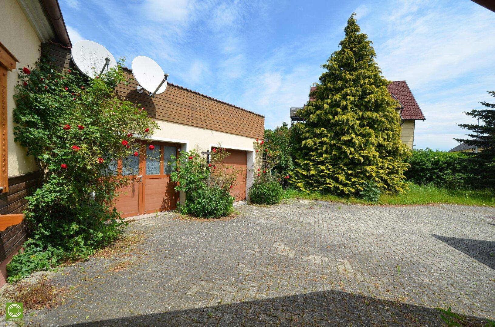 Gepflasterte Einfahrt mit Garagengebäude, umgeben von Rosenbüschen und hohen Bäumen.