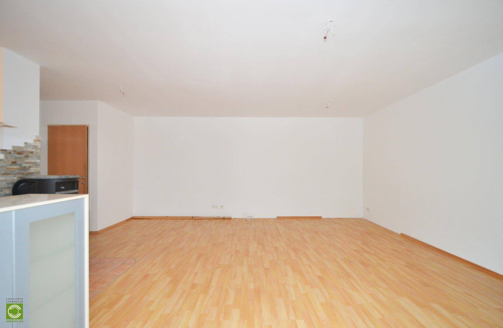 Heller, leerer Wohnbereich mit Laminatboden und Blick auf den Küchenbereich.
