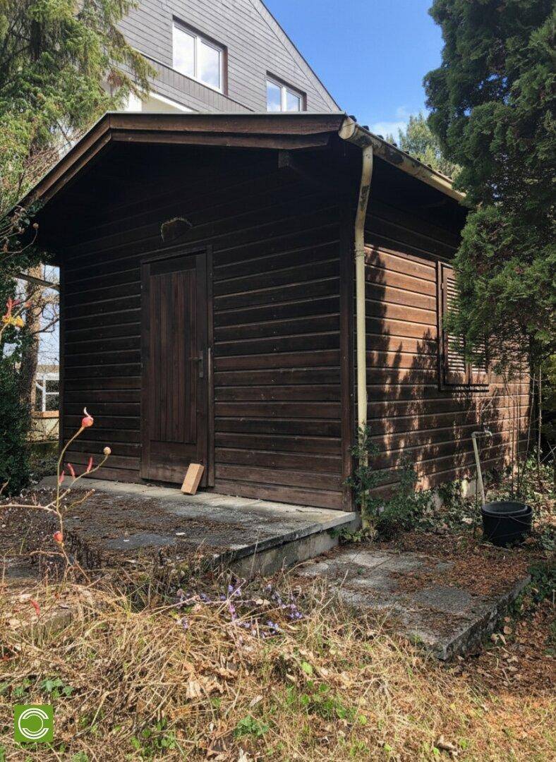 Rustikaler Holzschuppen mit einer kleinen Veranda und einer Holztür, umgeben von Vegetation.