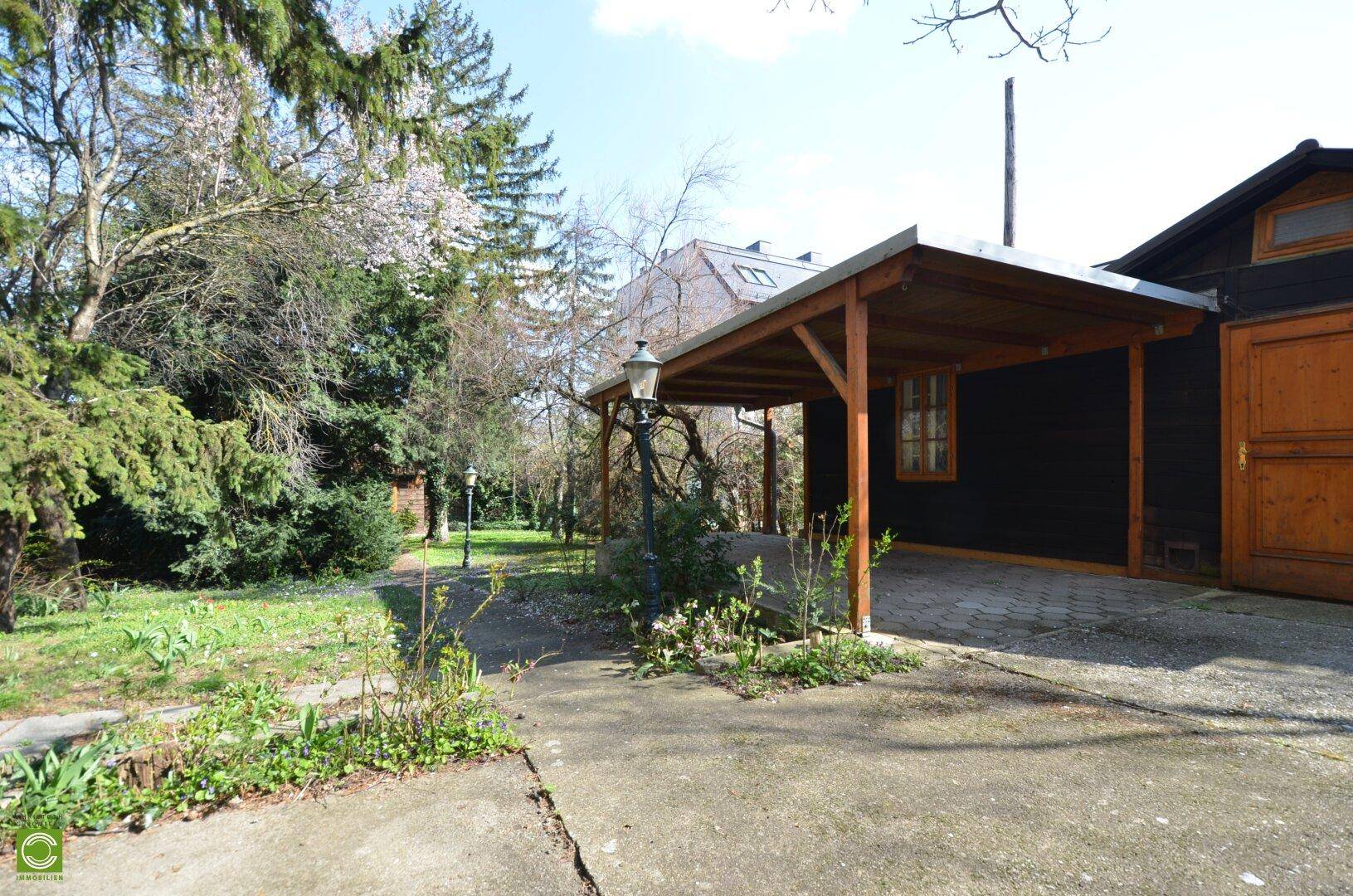 Großer Gartenbereich mit einem Holzcarport und einem kleinen Schuppen unter hohen Bäumen.