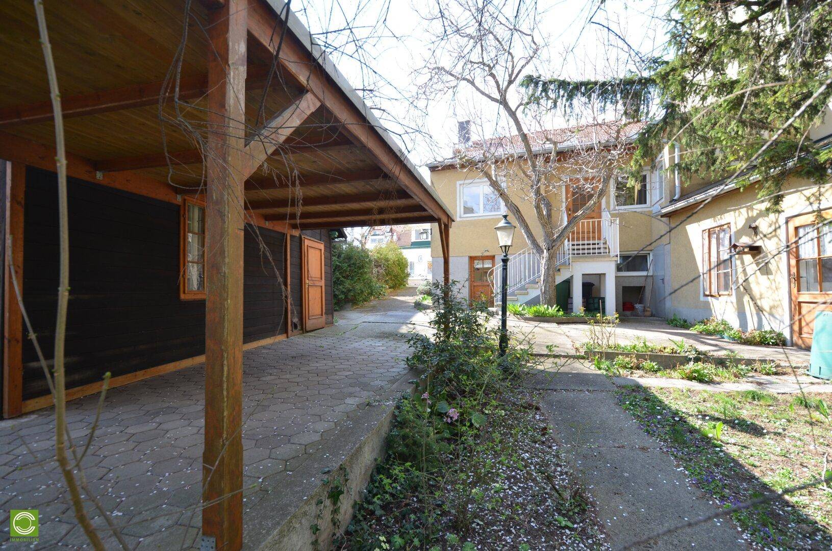 Blick auf die Rückseite des Hauses mit Carport, gepflastertem Weg und üppiger Vegetation.