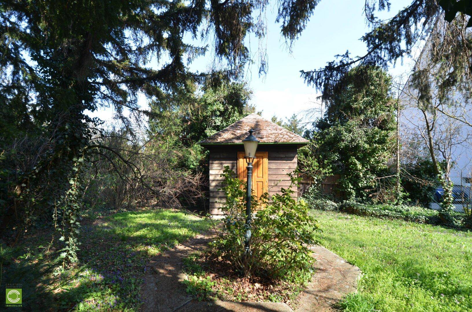 Kleiner Holzschuppen mit Laterne in einem weitläufigen, grünen Gartenbereich mit Bäumen.