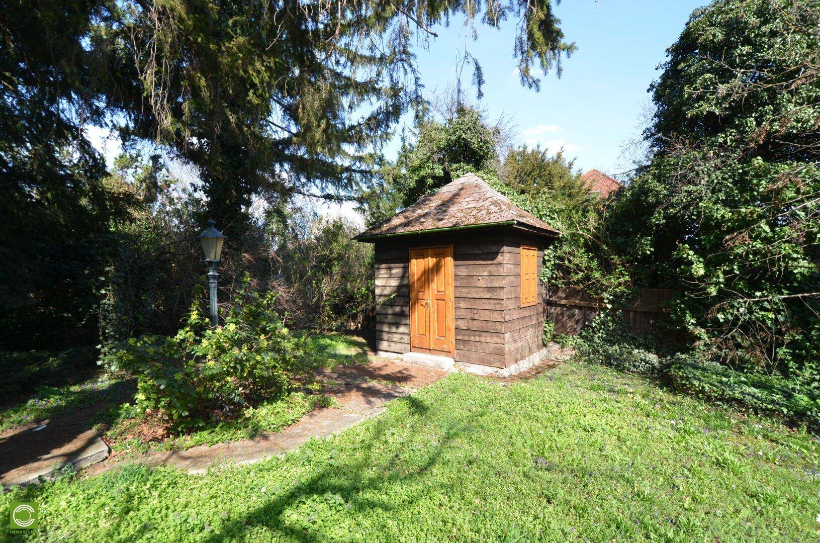 Rustikaler Holzschuppen mit braunen Türen, umgeben von grünem Rasen und Bäumen.