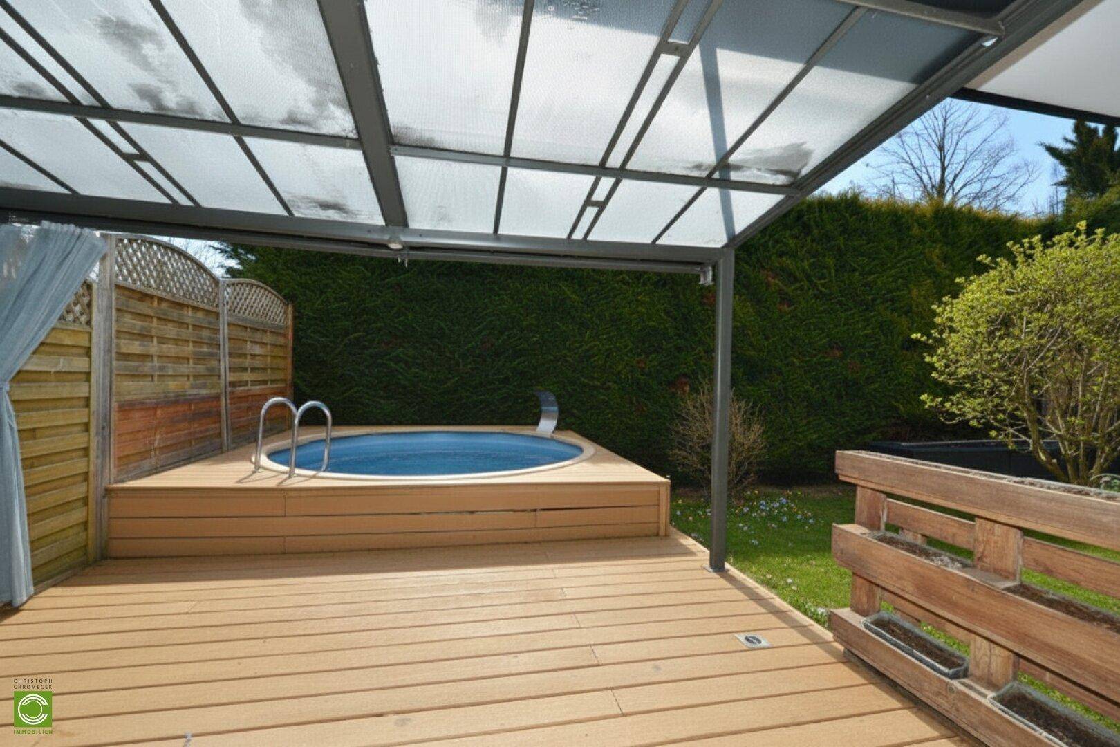 Überdachter Terrassenbereich mit Holzdeck und eingelassenem Whirlpool, umgeben von einer grünen Hecke.