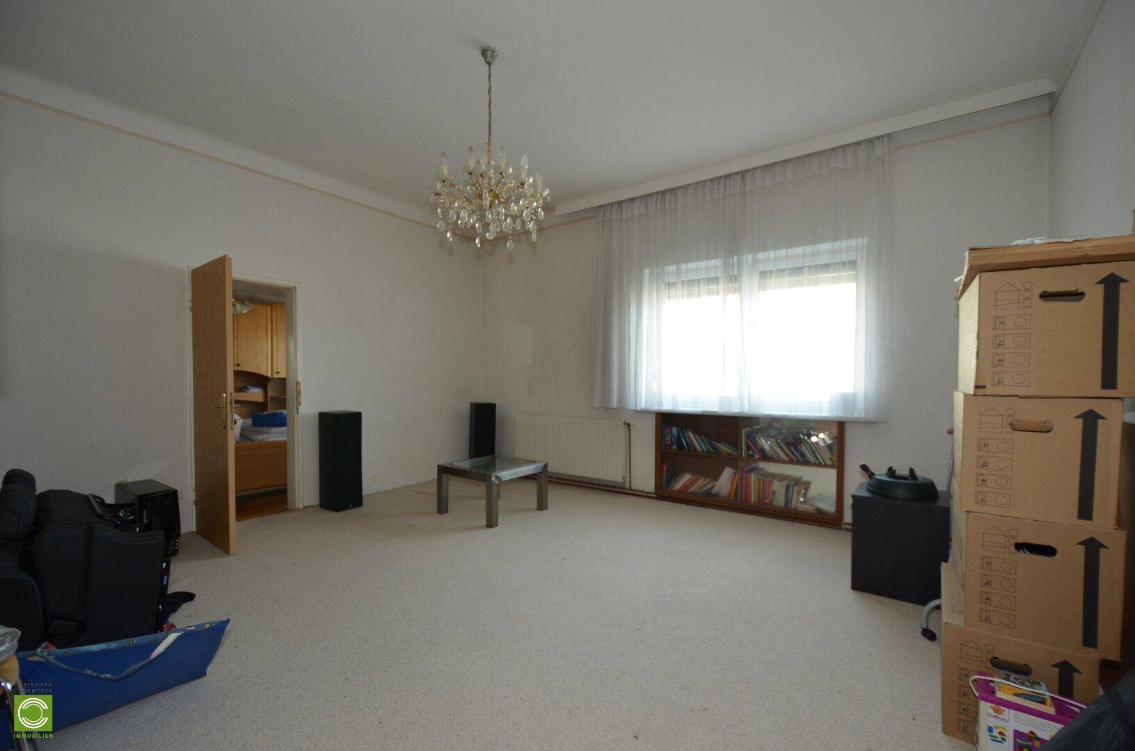 Geräumiges Wohnzimmer mit Teppichboden, Kronleuchter und einem großen Fenster, das viel Licht hereinlässt.
