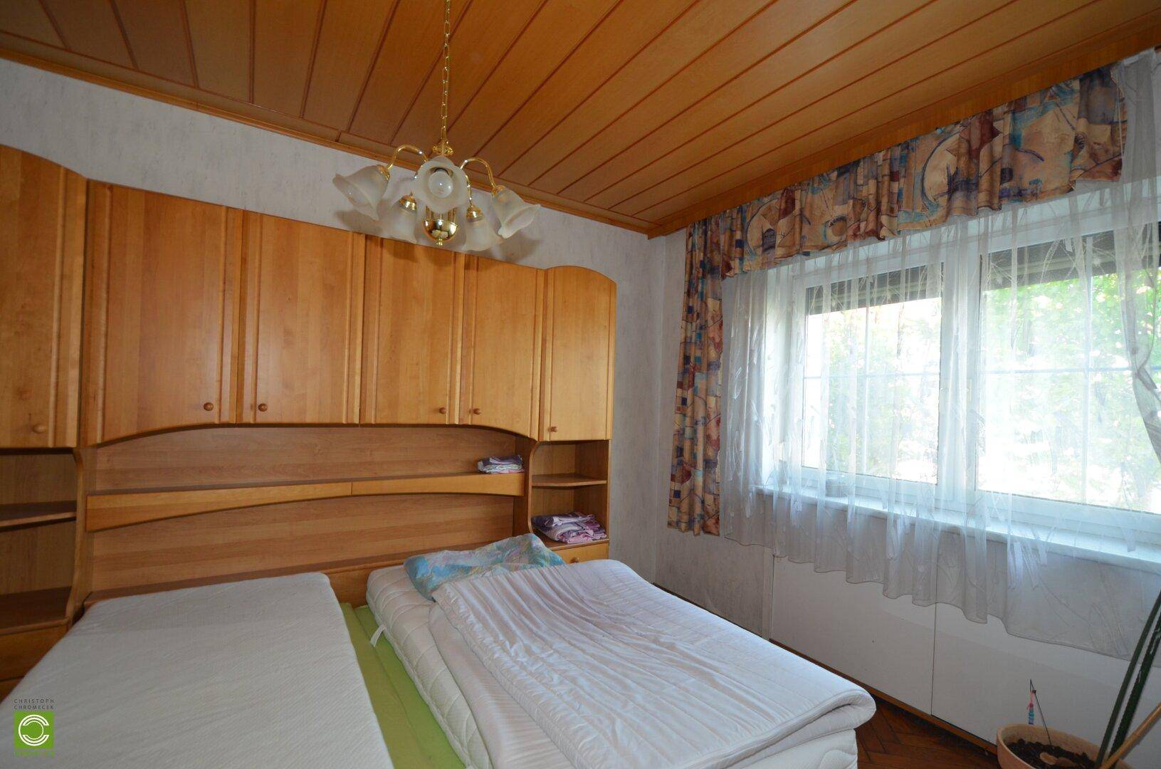 Schlafzimmer mit Holzdecke, Holzboden und einem großen Fenster, das den Raum erhellt.