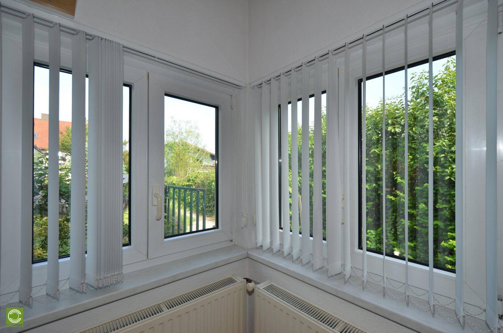Fensterecke mit vertikalen Jalousien und Heizkörper, bietet Blick ins Grüne.