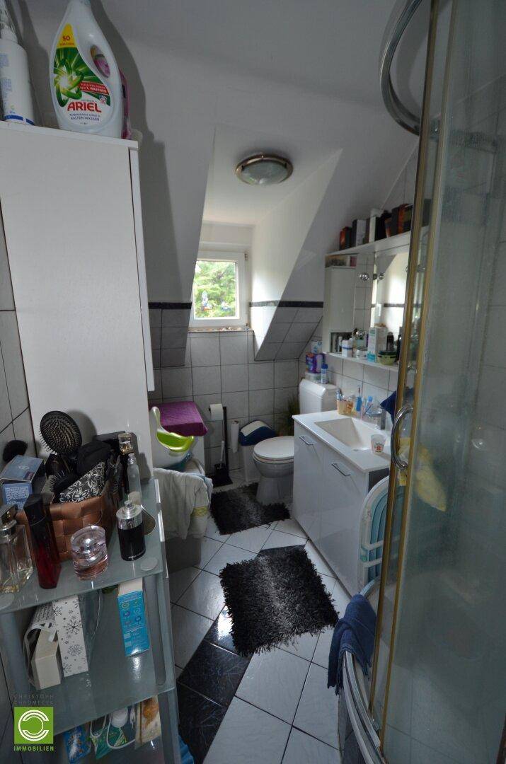Dachgeschoss-Badezimmer mit Dusche, WC, Waschbecken und Fenster, teilweise gefliest.