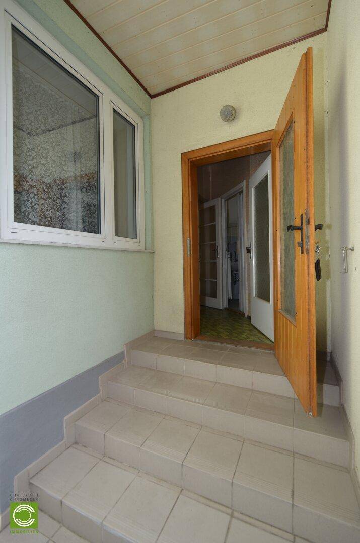 Eingangsbereich mit gefliesten Stufen, Fenster und Holztür, führt ins Innere des Hauses.