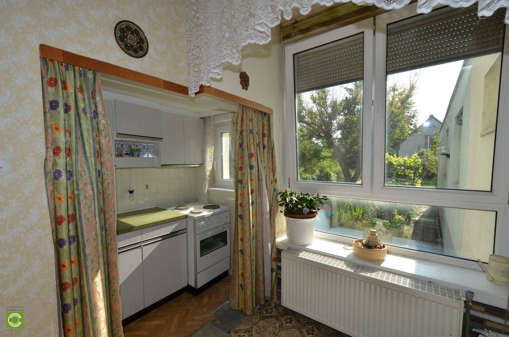 Kleine Küche mit Vorhängen, Fenster und Blick in den Garten, funktional eingerichtet.