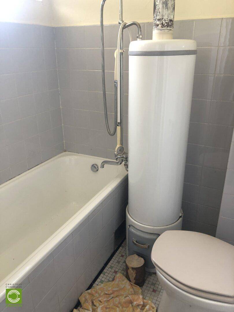 Badezimmer mit Badewanne, WC und Boiler, hell gefliest und mit Duschvorrichtung.