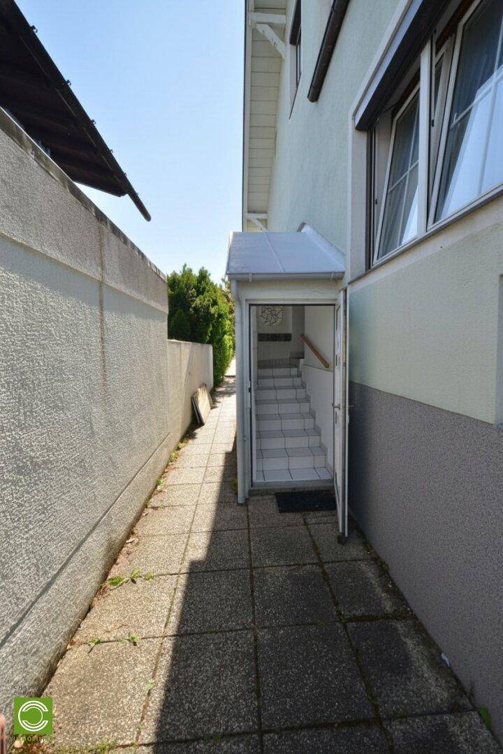 Schmaler Zugangsweg entlang der Hauswand mit Treppenabgang zu einem Nebeneingang.