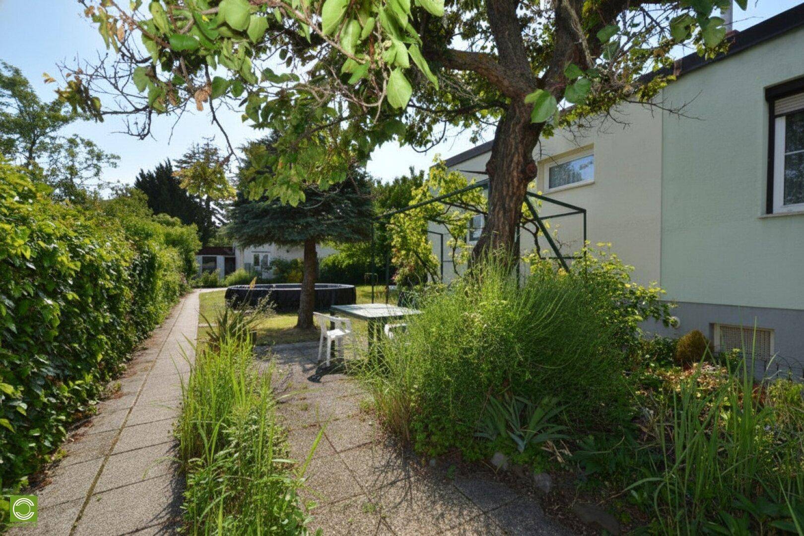 Langer Gartenweg mit üppiger Bepflanzung und einem Sitzbereich unter einem Baum.