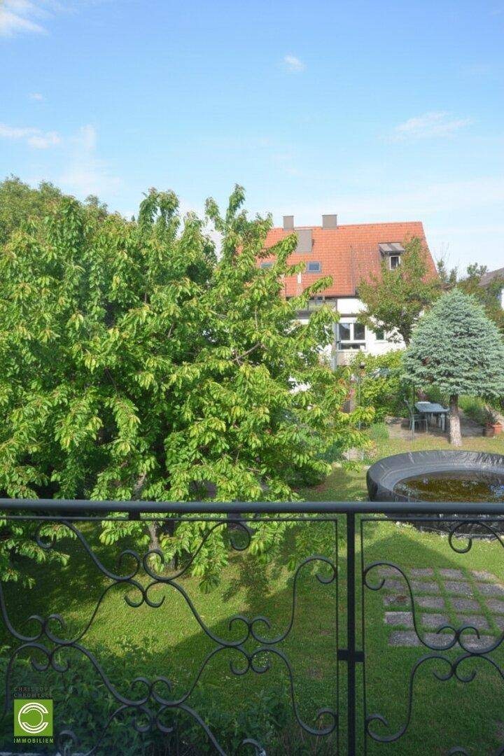 Blick vom Balkon auf den grünen Garten und das Nachbarhaus mit rotem Dach.
