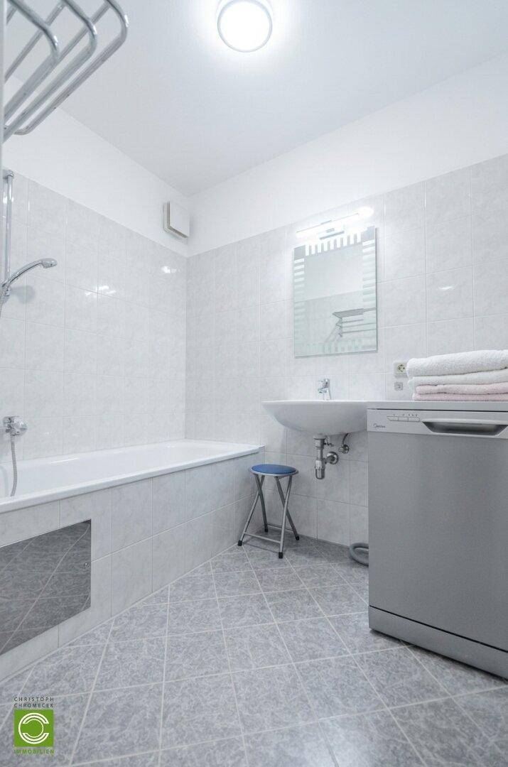 Badezimmer mit weißen Fliesen, Badewanne mit Duschfunktion, Waschbecken und Waschmaschine.