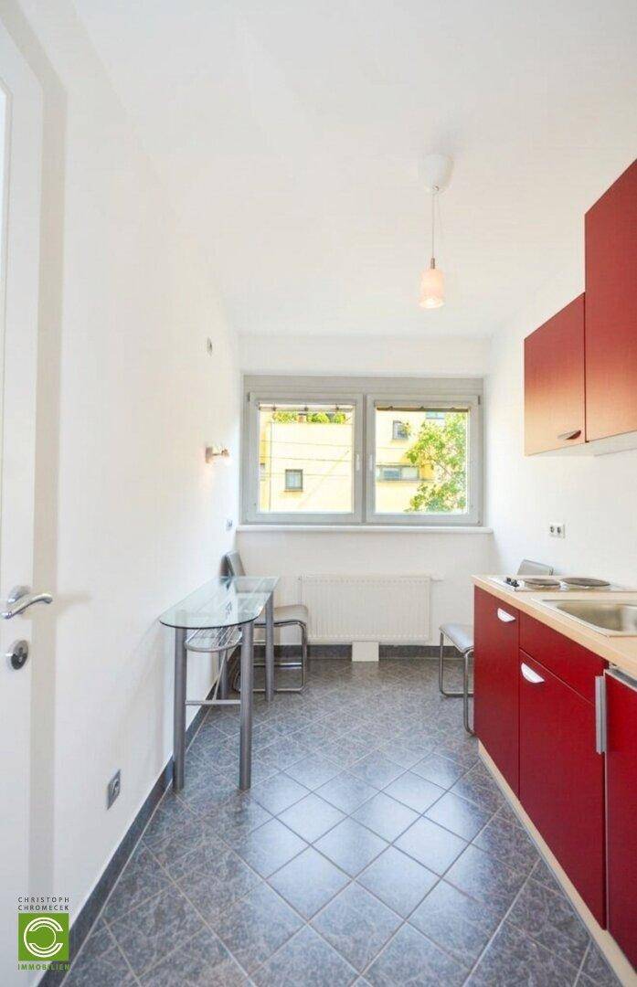 Kleine Küchenzeile mit roten Schränken, Fliesenboden und einem Esstisch am Fenster.