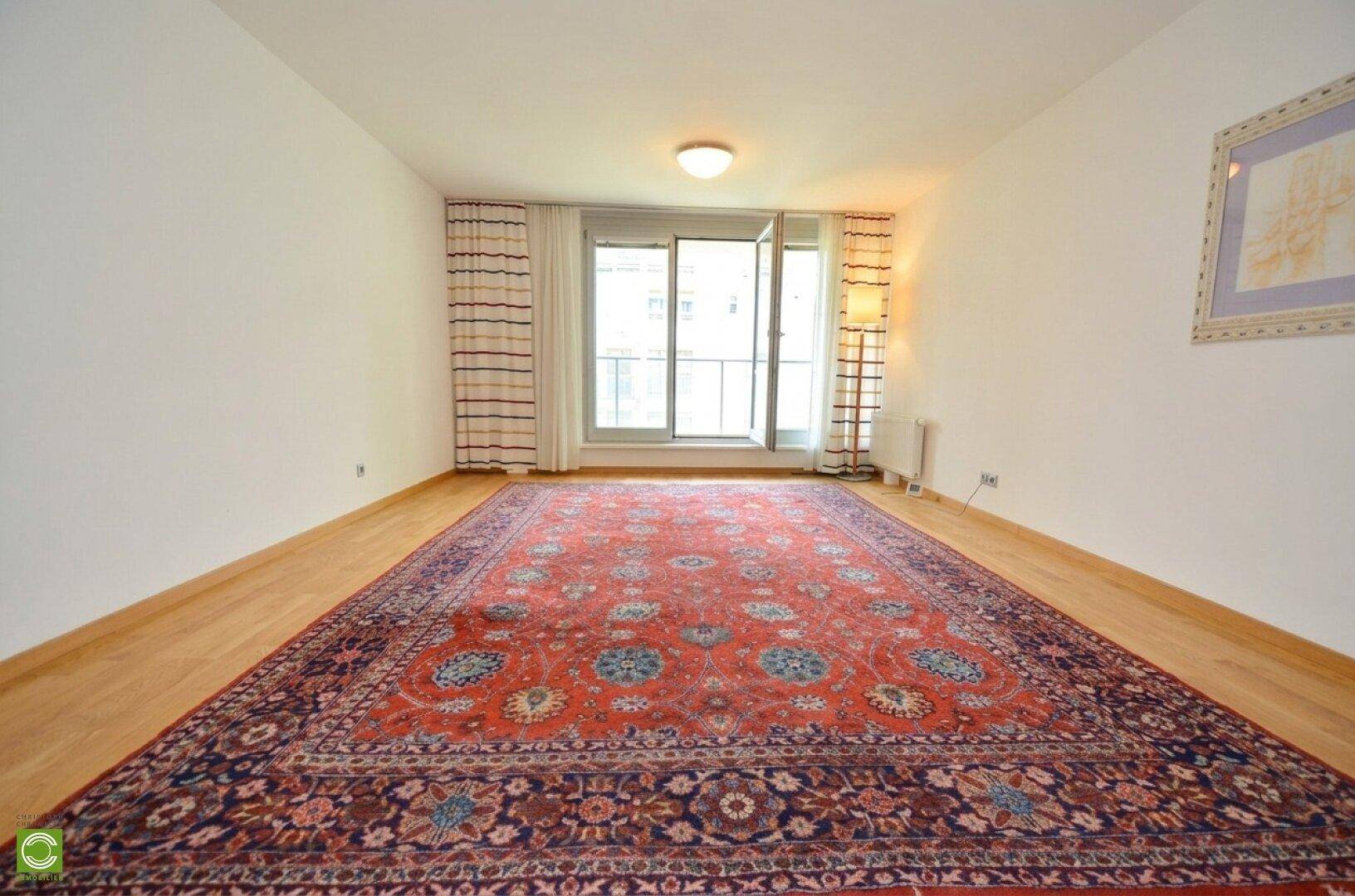Geräumiges Wohnzimmer mit Holzfußboden, großem Orientteppich und Zugang zum Balkon.