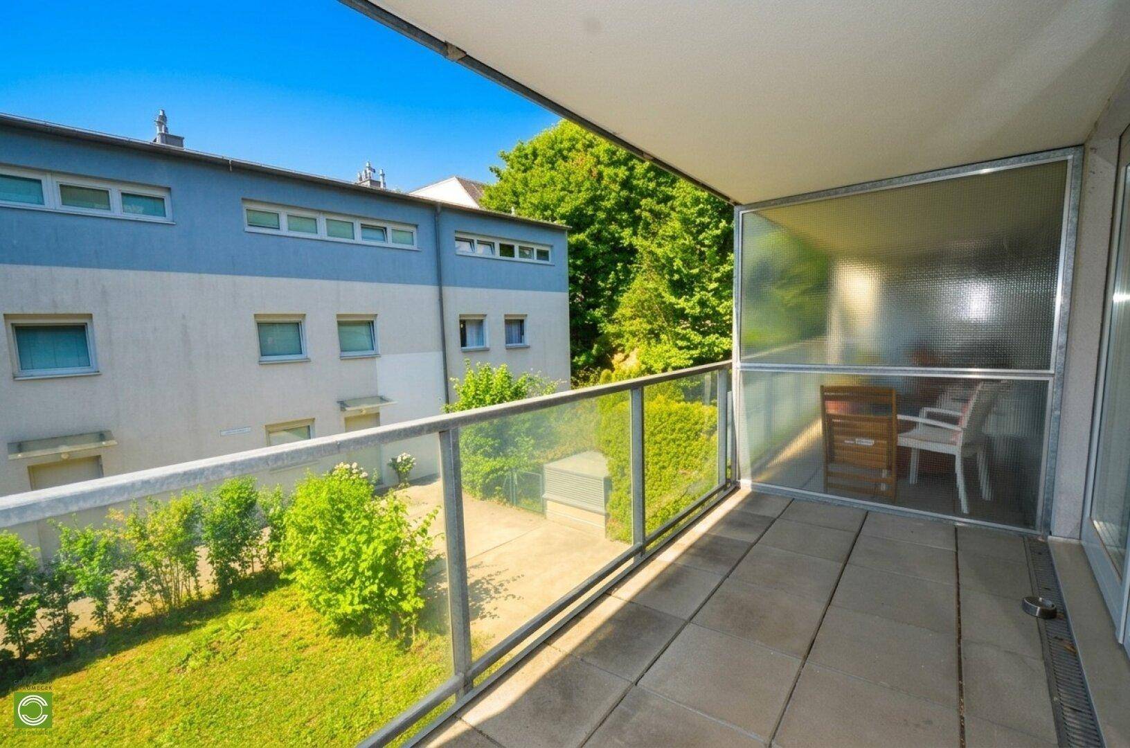 Heller Balkon mit Fliesenboden, Glasgeländer und Blick auf den grünen Innenhof.