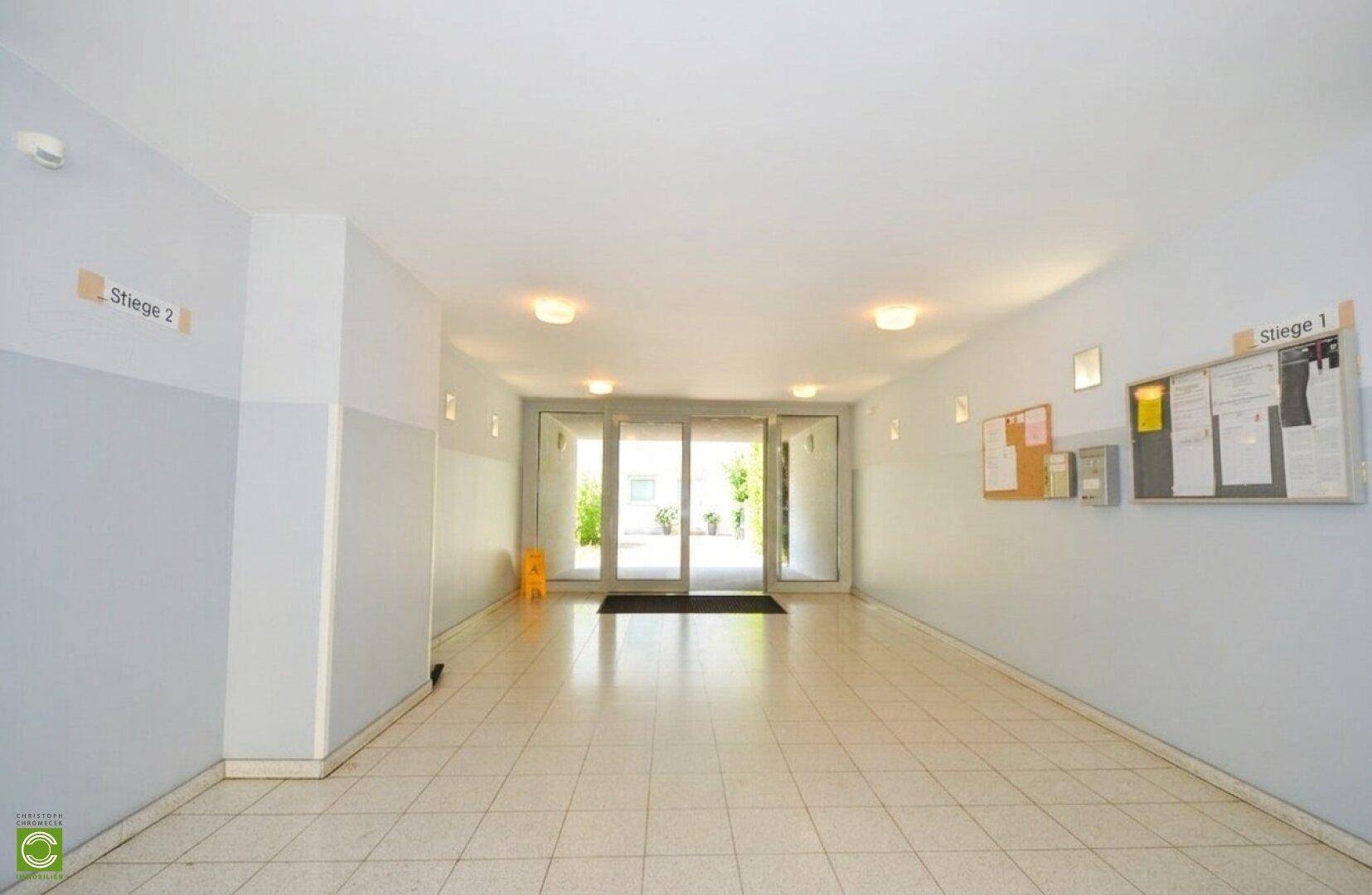 Heller Eingangsbereich des Gebäudes mit Fliesenboden und Glastüren zum Außenbereich.