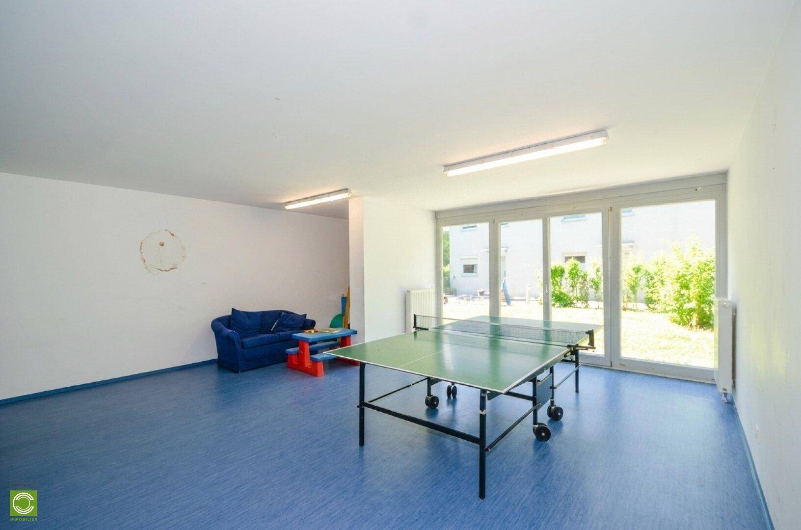 Gemeinschaftsraum mit Tischtennisplatte, blauem Boden und großen Fenstern zum Garten.