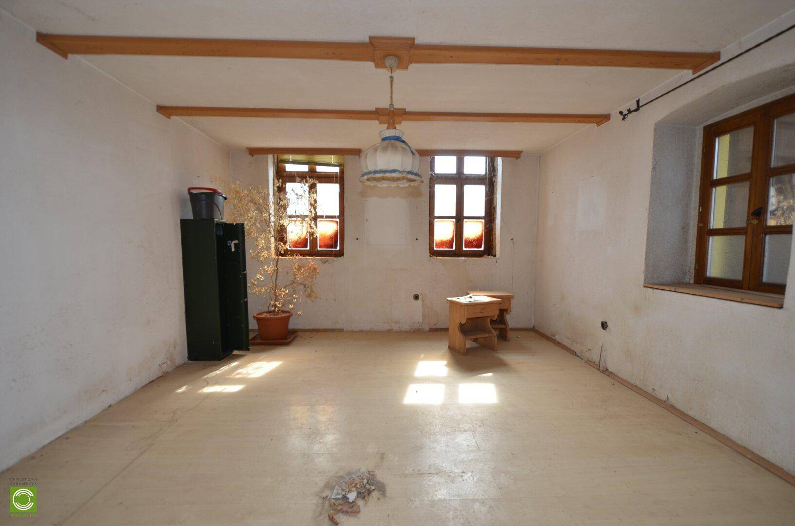 Leerer Raum mit sichtbaren Holzbalken an der Decke und zwei Fenstern, bereit zur Renovierung.