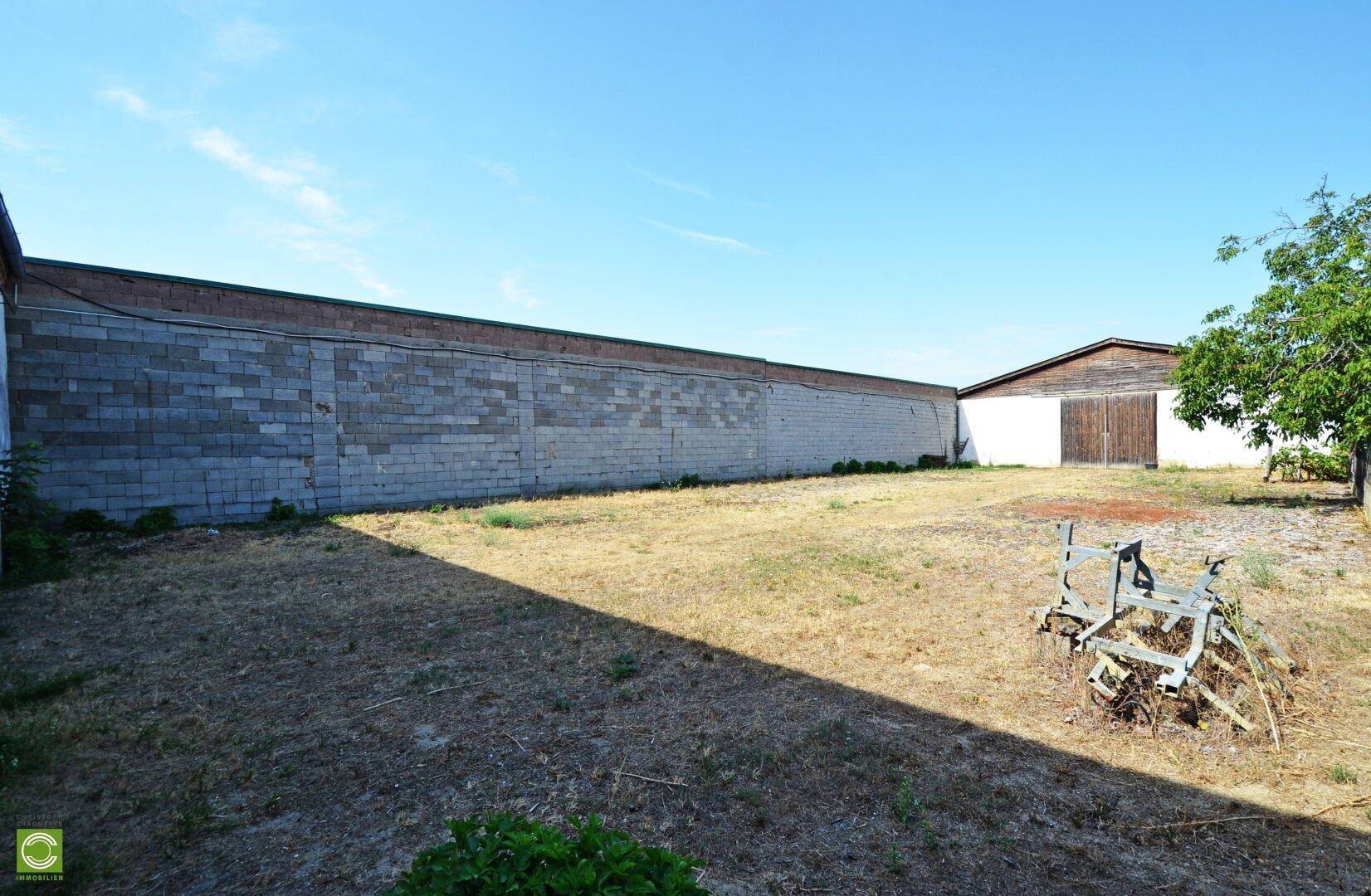 Großes, trockenes Gartengrundstück mit langer Mauer und einem älteren Gebäude unter blauem Himmel.