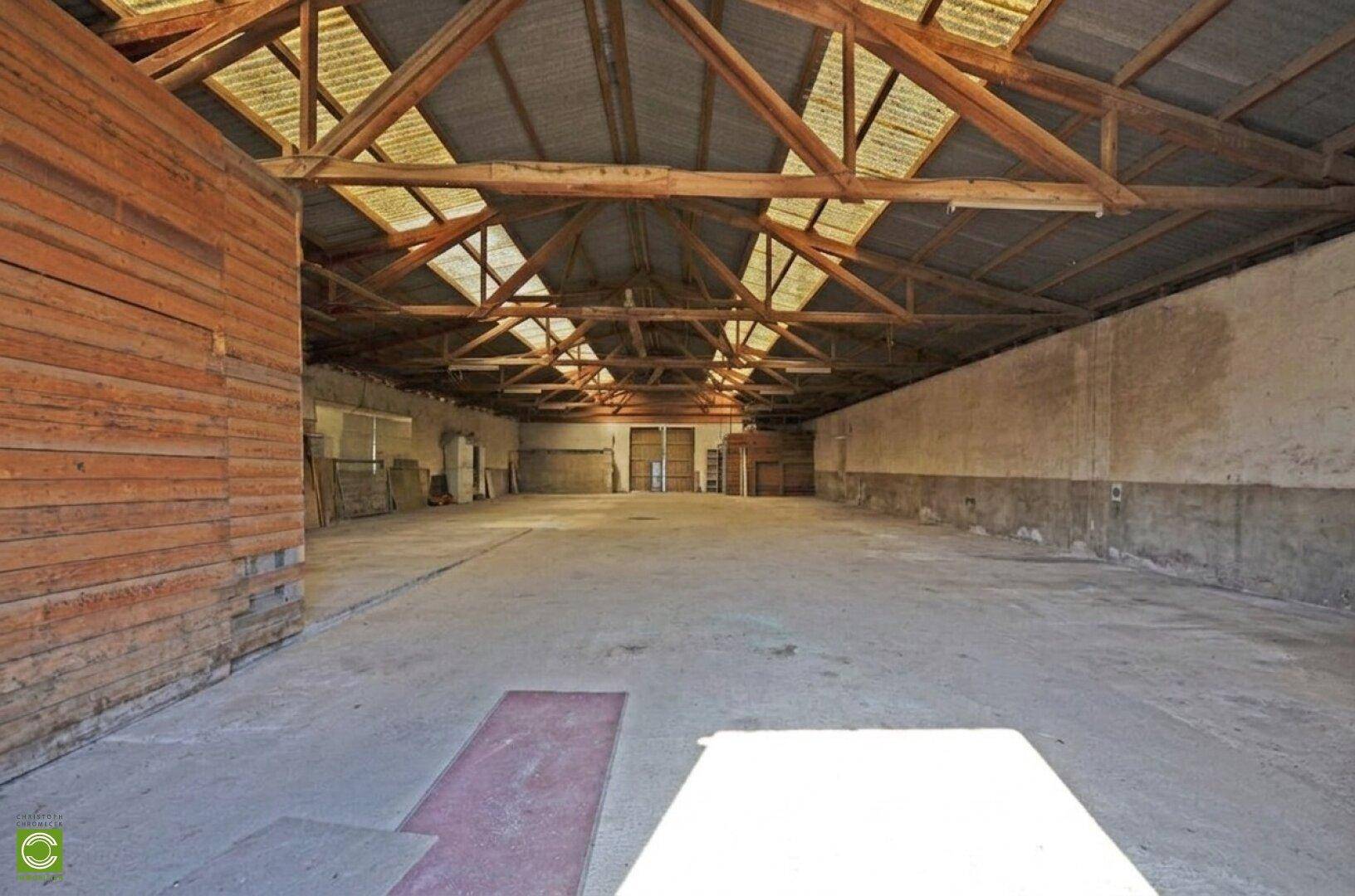 Große, leere Halle mit Betonboden und sichtbarer Holzkonstruktion des Daches, beleuchtet durch Oberlichter.