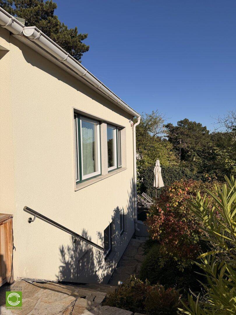 Seitenansicht des Hauses mit Fenstern, einer Treppe und einem gepflegten Gartenbereich mit Sonnenschirm.