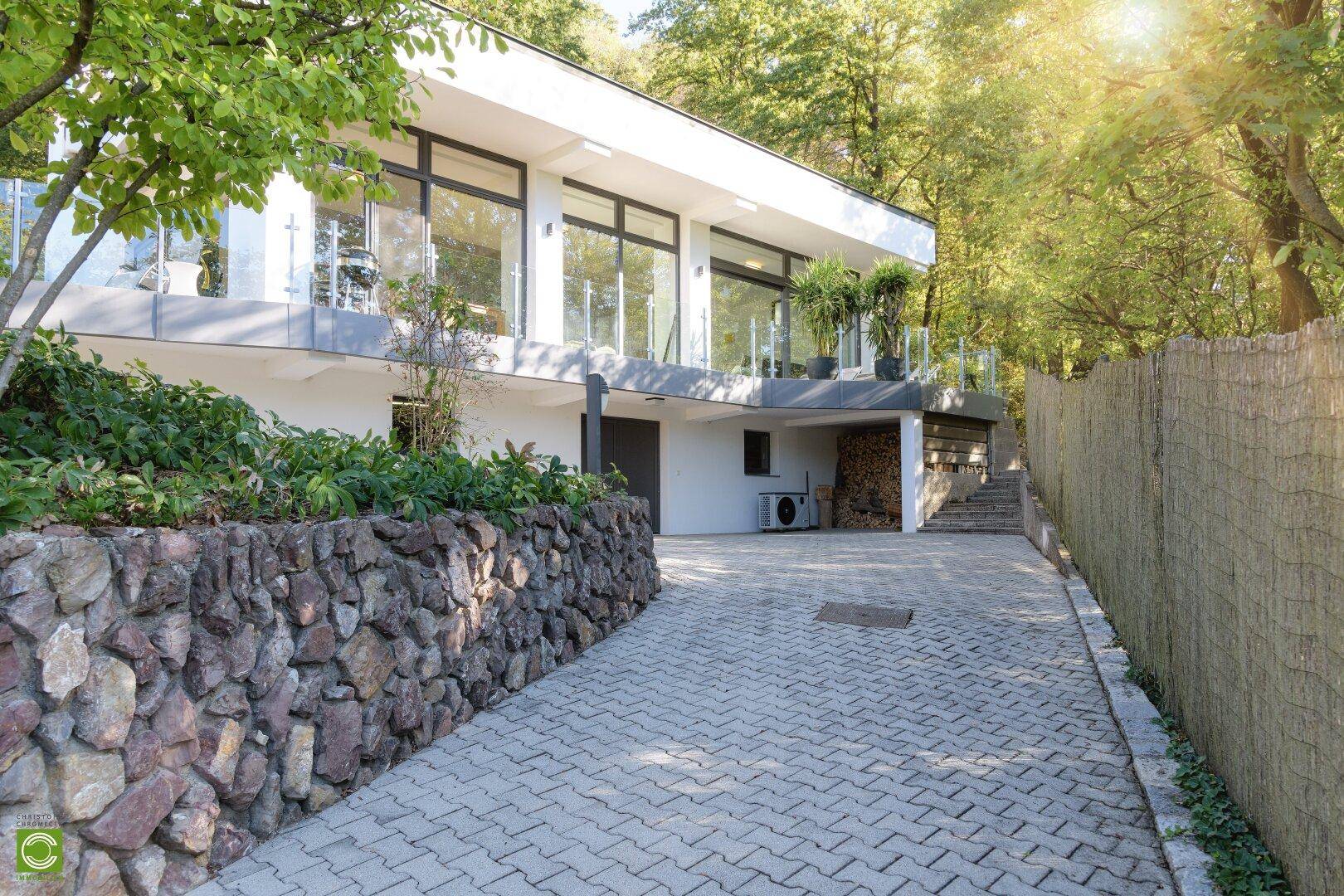 Moderne Hausfassade mit großen Fenstern, Auffahrt und einer Natursteinmauer im Grünen.